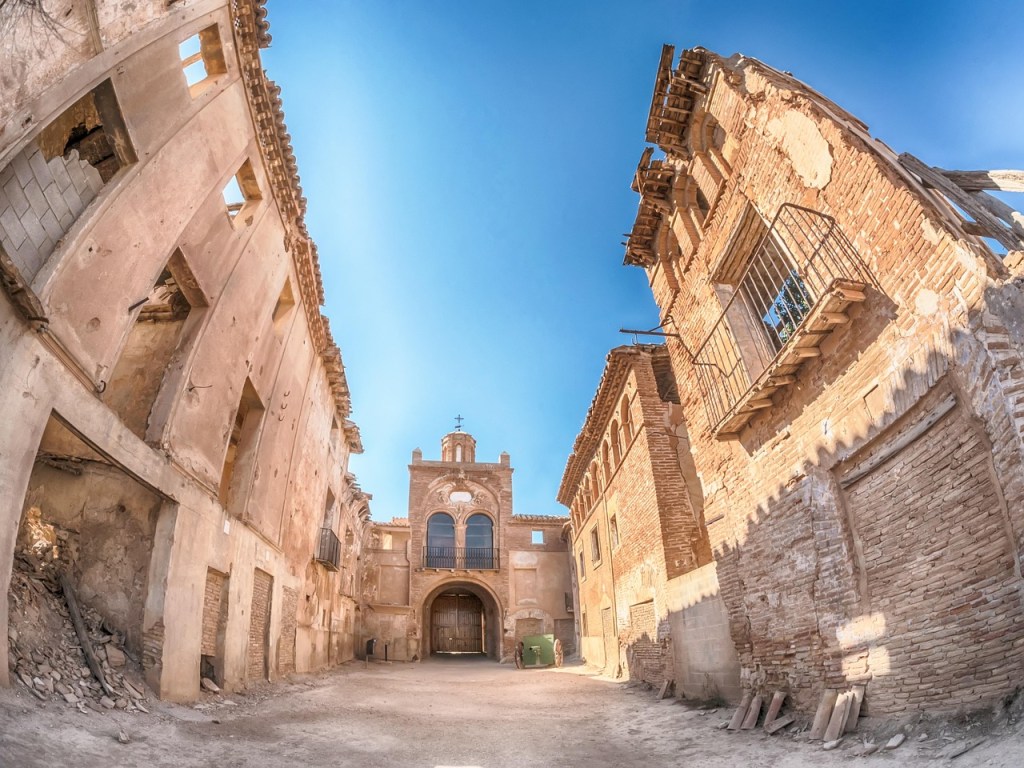 View of a dilapidated building complex in Syria with partially collapsed walls and clear blue sky above, suggesting remnants of a historical structure, possibly affected by conflict.